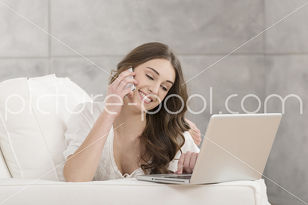 junge Frau mit Laptop liegt auf Sofa und telefoniert 