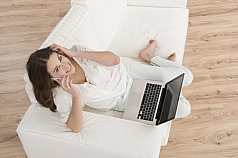 junge Frau mit Laptop sitzt telefonierend auf dem Sofa 